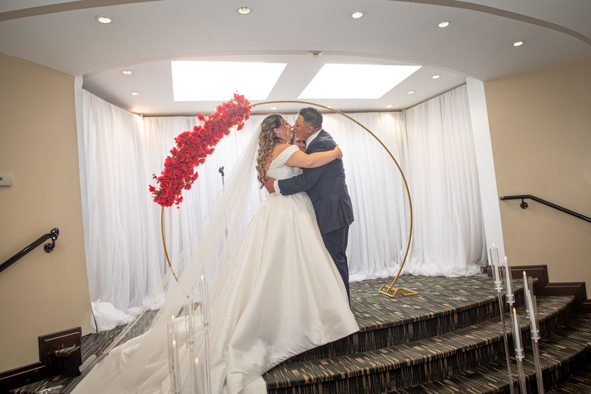 A newlywed couple kisses on an indoor staircase under a golden arch decorated with red flowers and cascading vines.