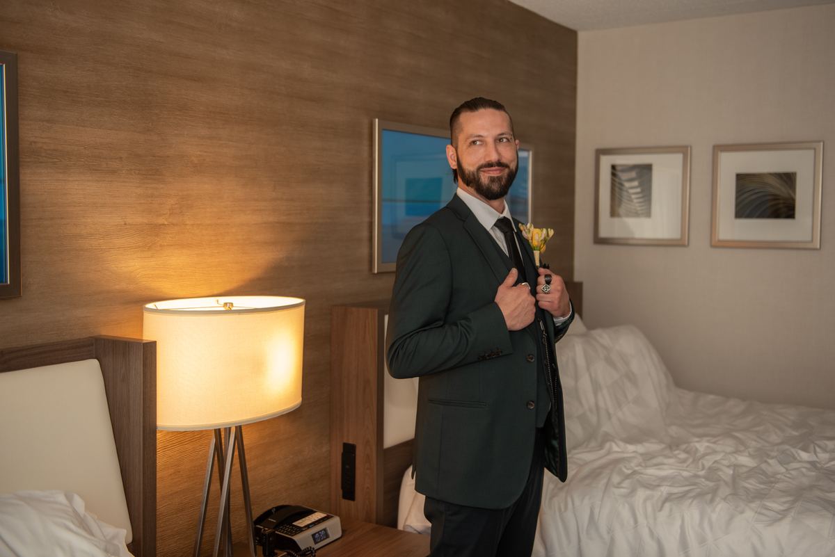 A groom adjusts his suit jacket, holding a boutonnière in a warmly lit hotel room, highlighting his composed expression.