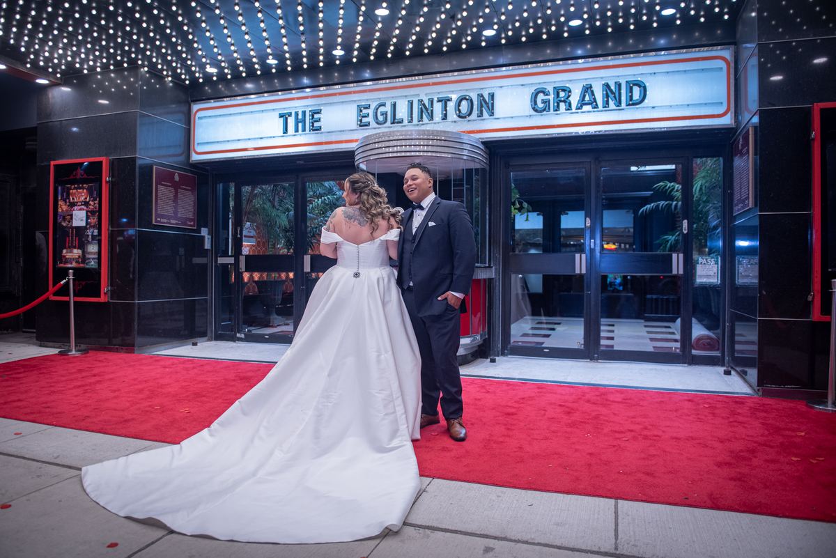 A laughing bride and groom stand on a red carpet in front of a theater's illuminated marquee, surrounded by string lights.