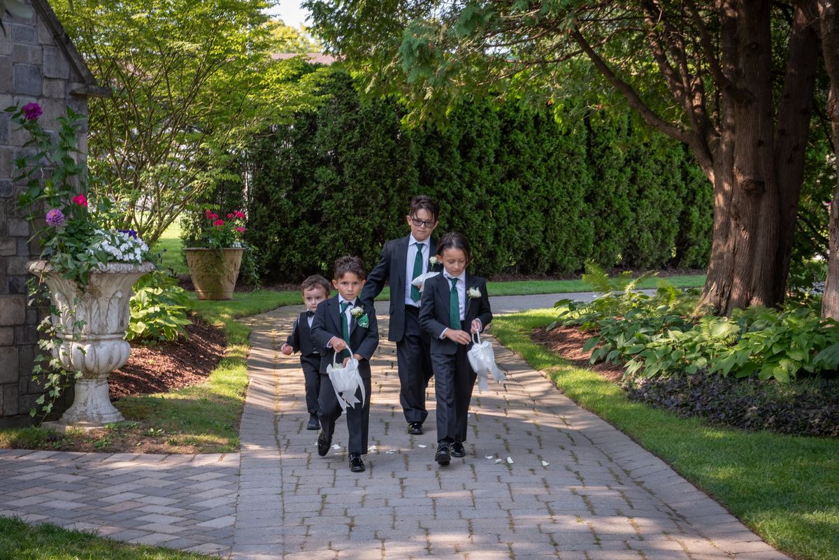 Four boys in dark suits and green ties walk on a stone path, scattering flower petals under sunlight filtering through trees.