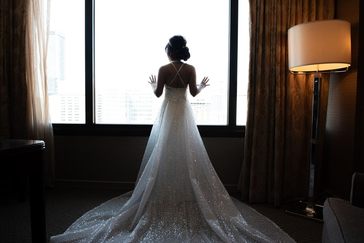 A bride in a sparkling gown stands by a window, gazing at a cityscape, illuminated by soft light from a floor lamp.