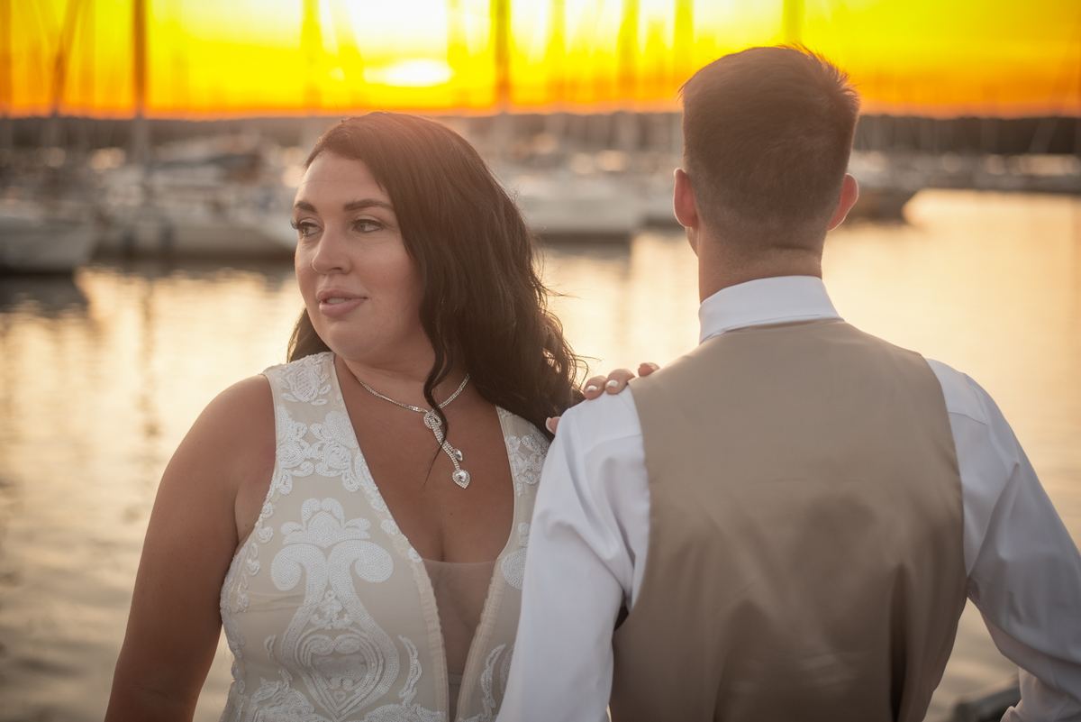 A bride and groom stand close at sunset, gazing tenderly as golden light reflects off water behind them, shoulder.