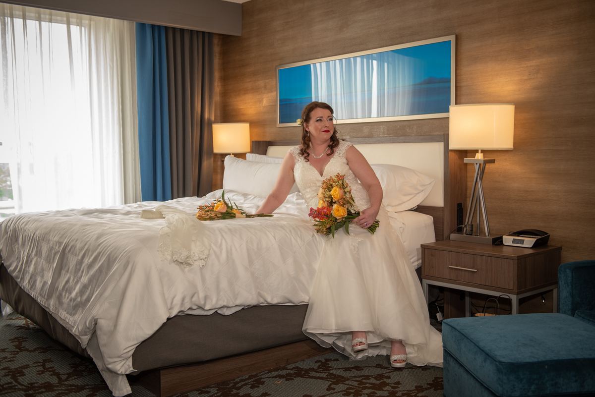 A bride in a white gown and yellow and orange flowers sits on a hotel bed, gazing thoughtfully at the camera.