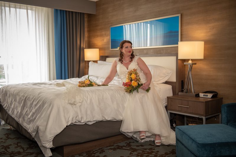 A bride in a white gown and yellow and orange flowers sits on a hotel bed, gazing thoughtfully at the camera.