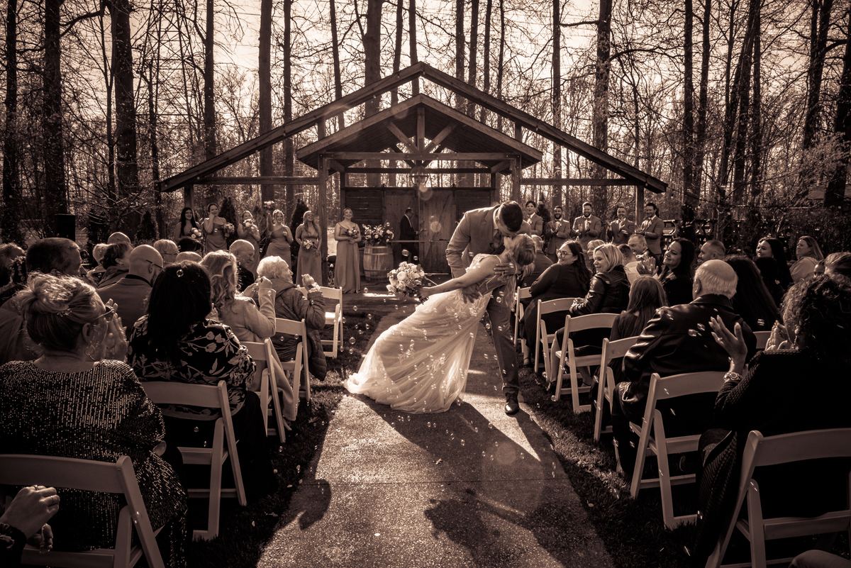 A bride and groom kiss at an outdoor ceremony, confetti drifting around them as sunlight filters through trees.