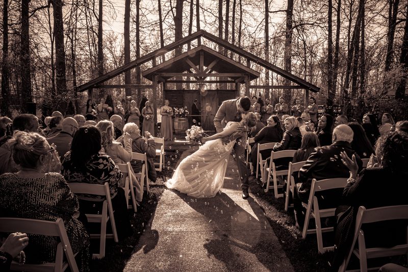 A bride and groom kiss at an outdoor ceremony, confetti drifting around them as sunlight filters through trees.