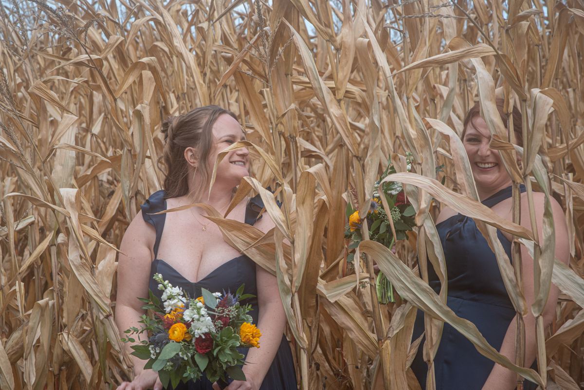Two women in dark navy dresses stand smiling in a dry cornfield with autumnal bouquets, celebrating.
