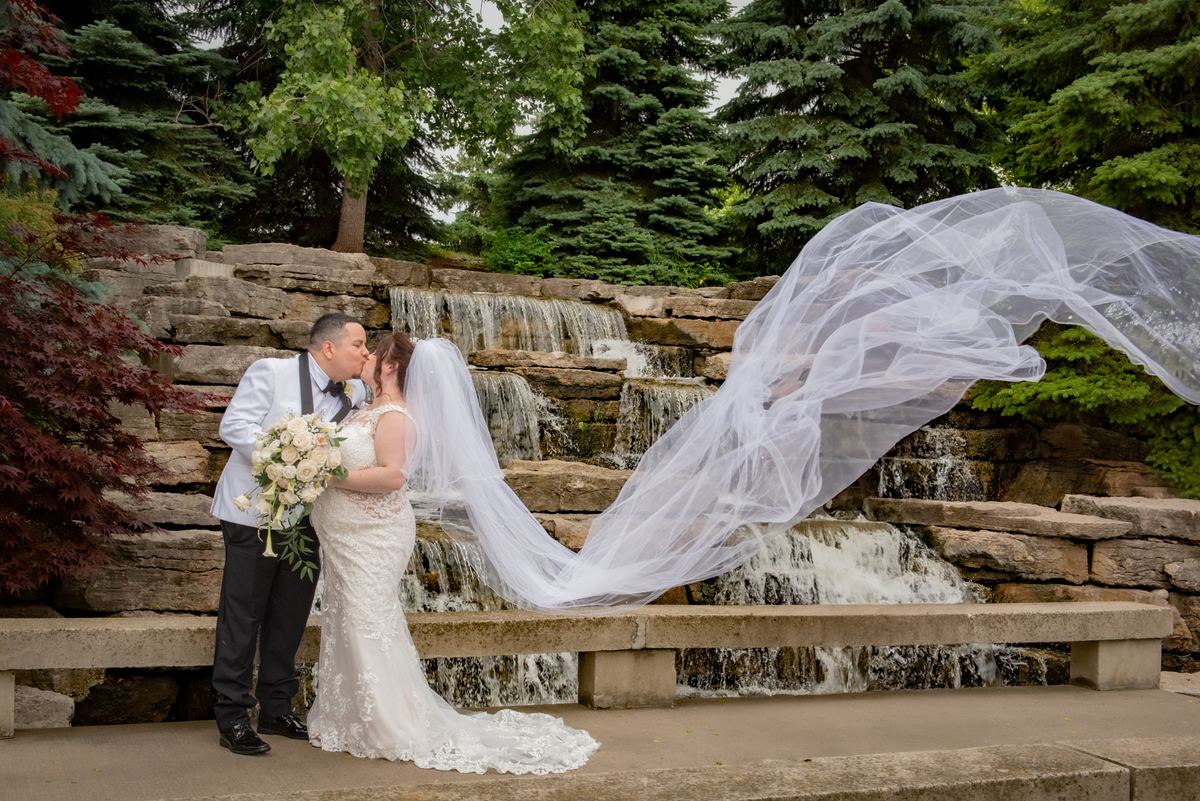 A bride and groom kiss in front of a stone waterfall, with her veil billowing in the wind among lush greenery.