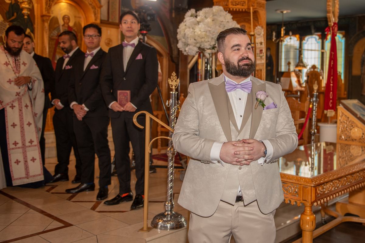 A groom in a light beige tuxedo stands with his back to an Orthodox priest and groomsmen in black suits.