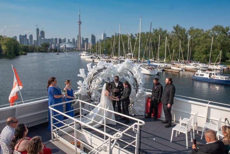 Bride and groom stand under floral arch on boat deck with Toronto skyline visible, surrounded by guests and sailboats.