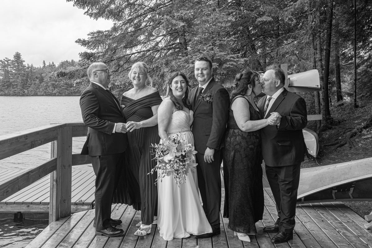 A joyful wedding party stands on a lakeside dock, smiling and embracing, with the bride and groom at center.