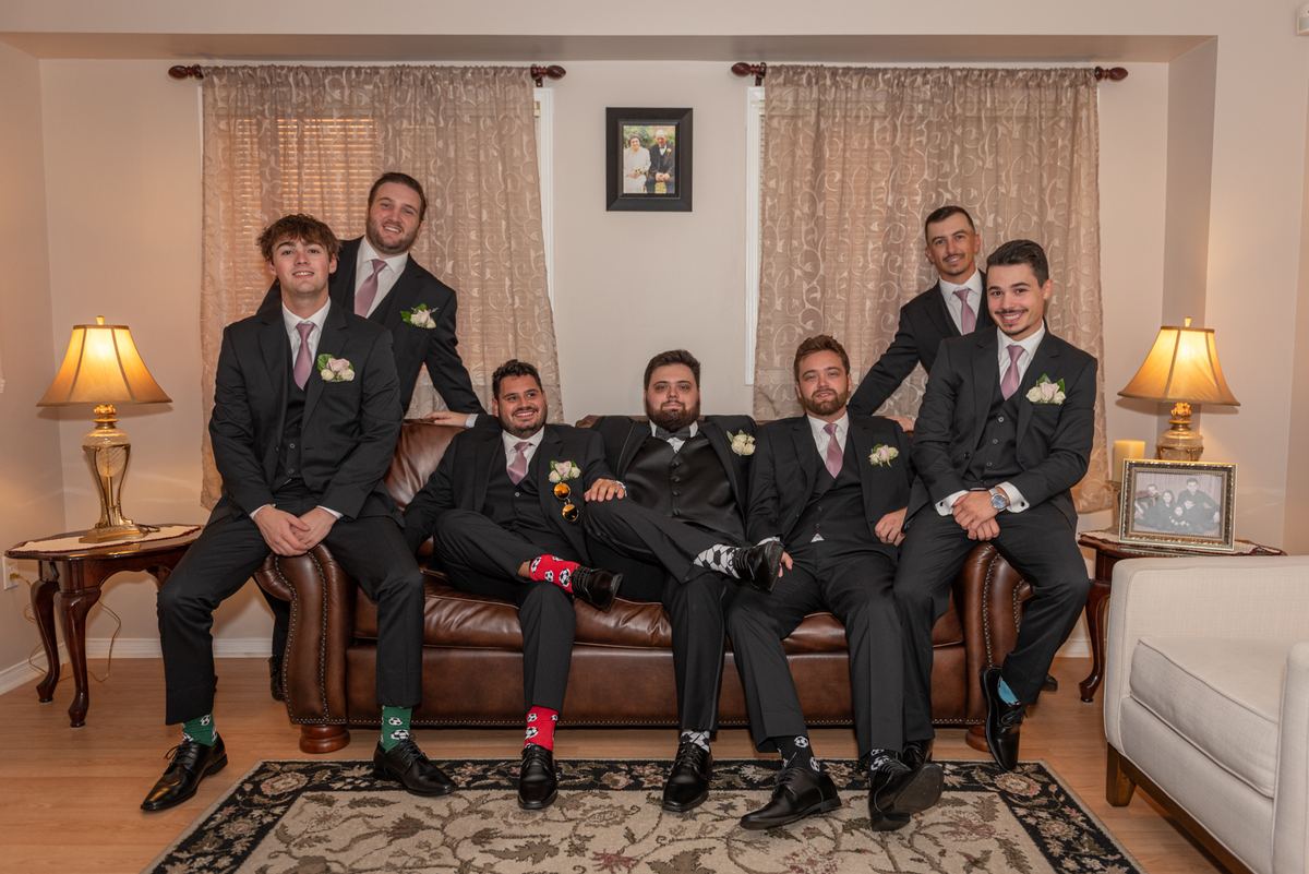 Seven groomsmen in black suits with colorful socks pose on a leather sofa in a warmly lit living room.