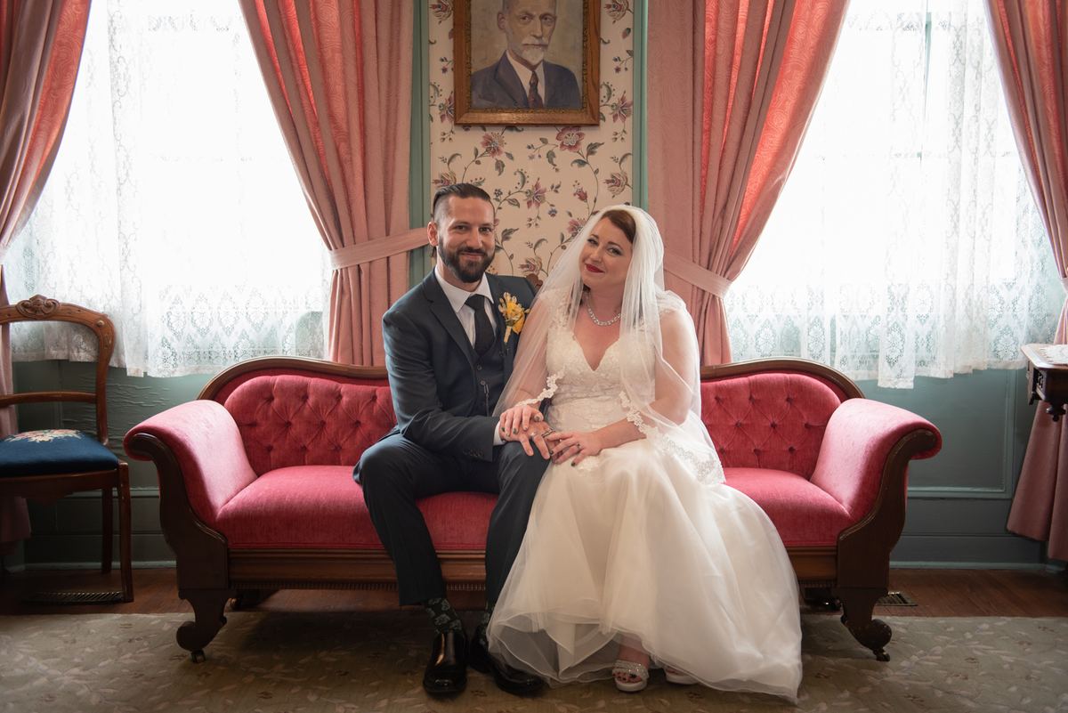 A newlywed couple in formal attire sit on a vintage red sofa, smiling in an ornate room with floral wallpaper.