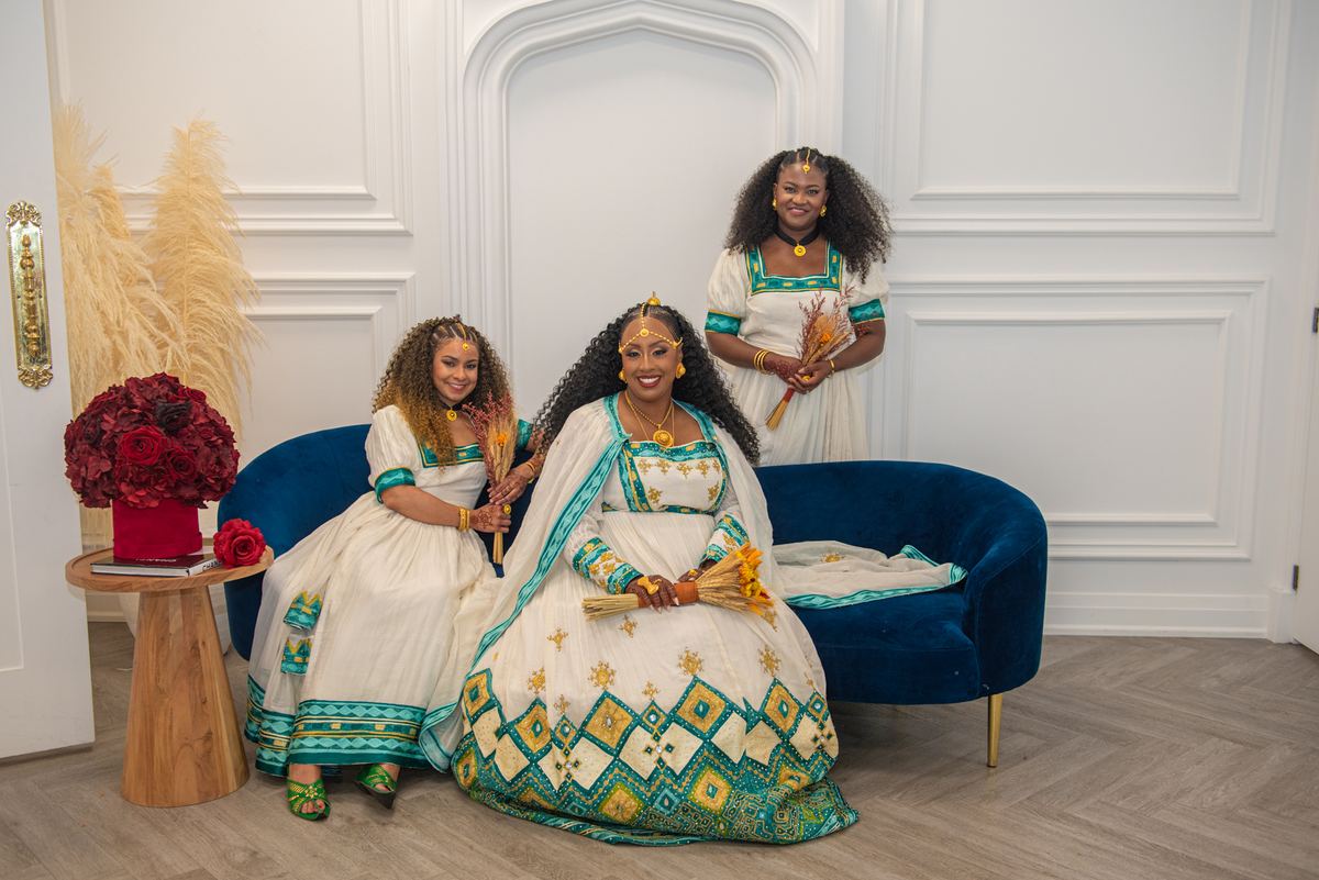 Three women in vibrant traditional African attire pose with dried wheat stalks in an elegantly decorated room, smiling.