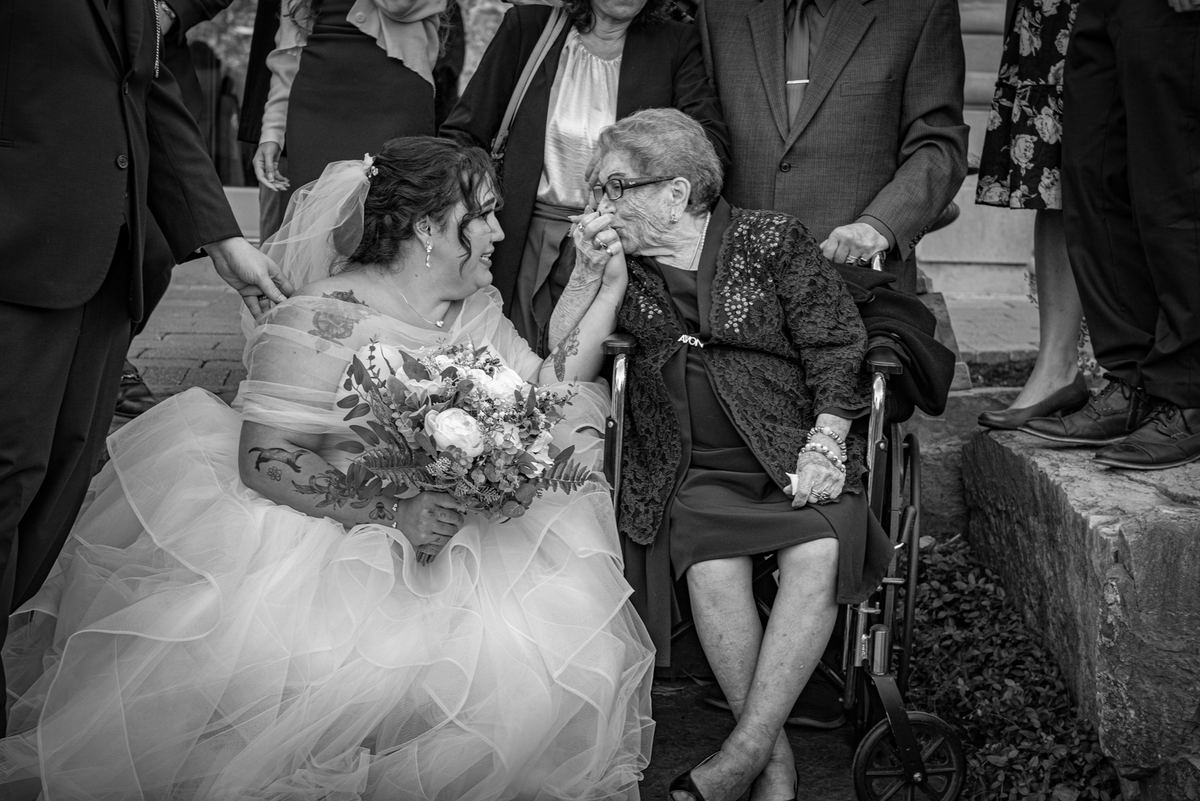 A bride leans in to touch an elderly woman's face in a tender moment at what seems like a wedding, illuminated by soft light.