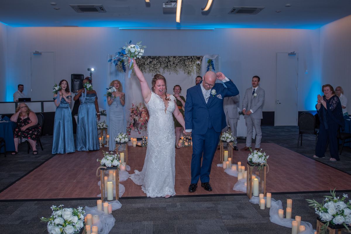 A bride and groom walk down a candlelit aisle, guests applauding and bridesmaids in blue gowns lining the path.