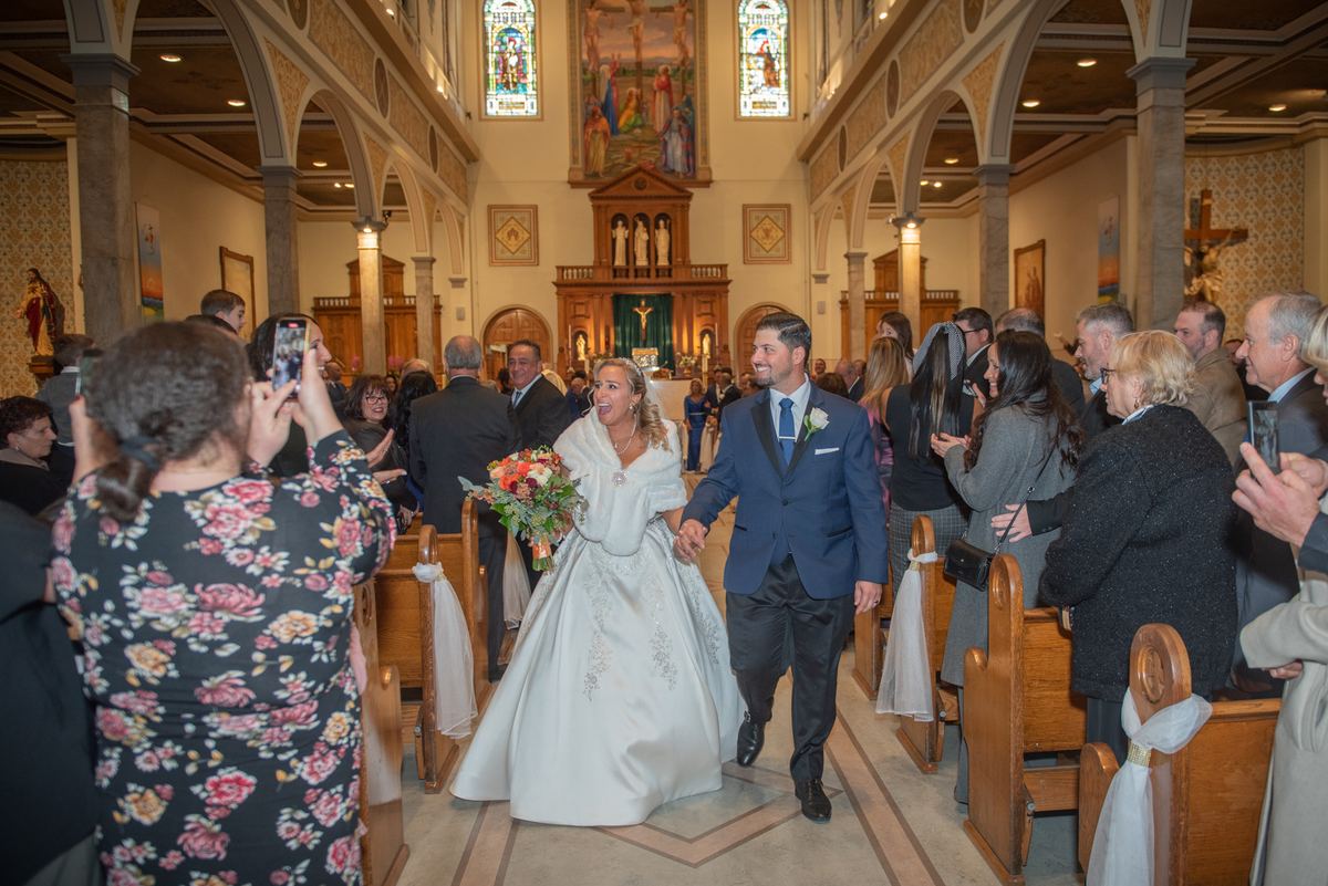 A smiling bride and groom walk down the aisle of a church, surrounded by guests taking photos on their phones.