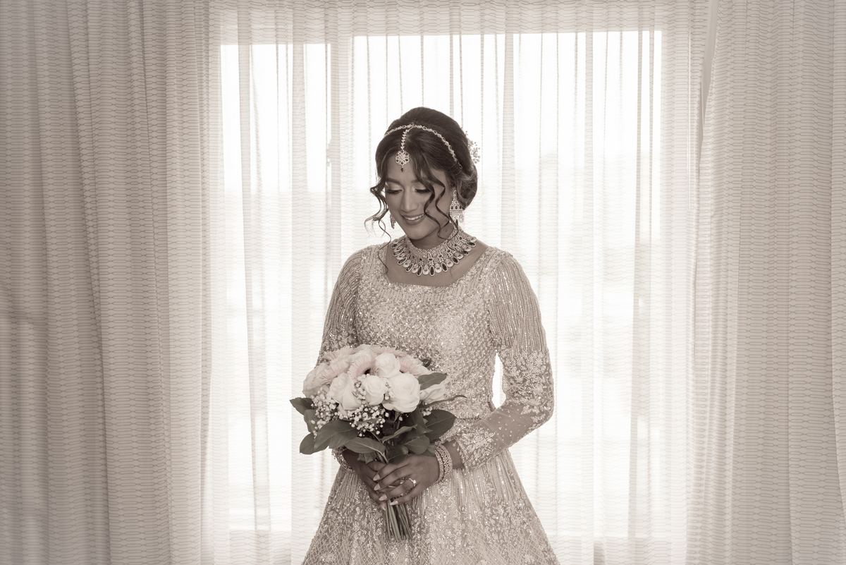 A bride in traditional attire and jewelry stands by sheer curtains, holding a bouquet with a serene expression.