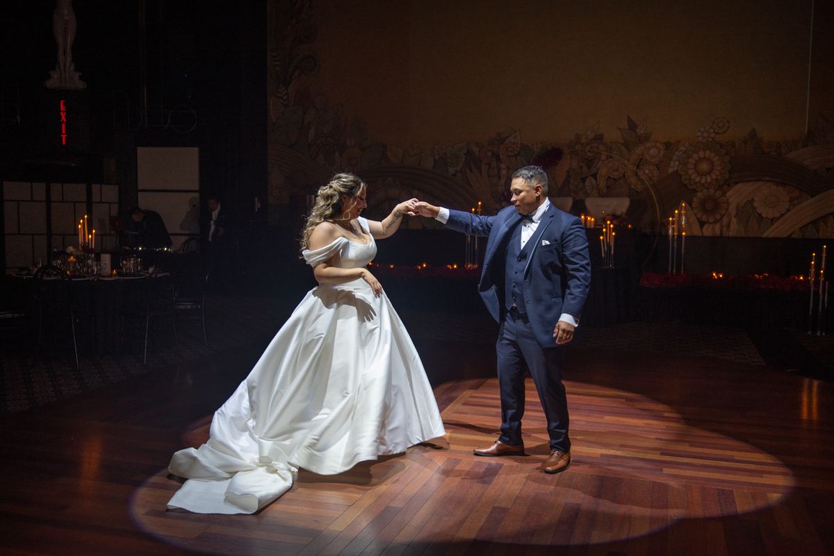 Bride and groom dance intimately under spotlight on wooden floor, hands clasped, in elegantly lit ballroom.