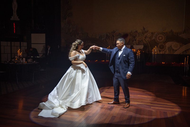 Bride and groom dance intimately under spotlight on wooden floor, hands clasped, in elegantly lit ballroom.