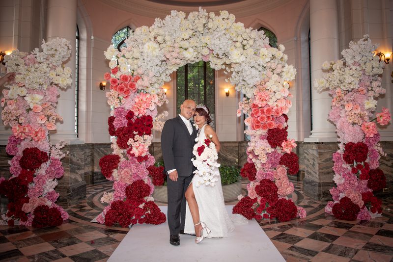A couple stands under a floral arch in an elegant hall with checkered flooring, illuminated by warm sconce light.