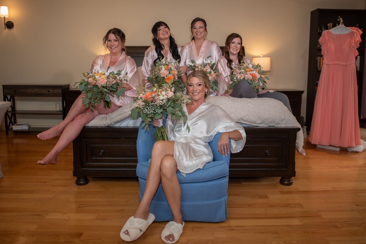 A bride and her bridesmaids in a warmly lit bedroom, all dressed in satin robes and holding floral bouquets.