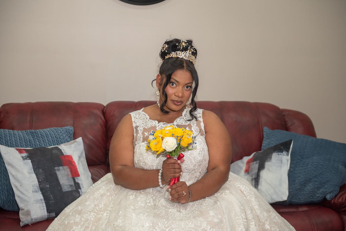 A bride in a white lace gown sits playfully on a red leather couch with a bouquet of yellow and white roses.