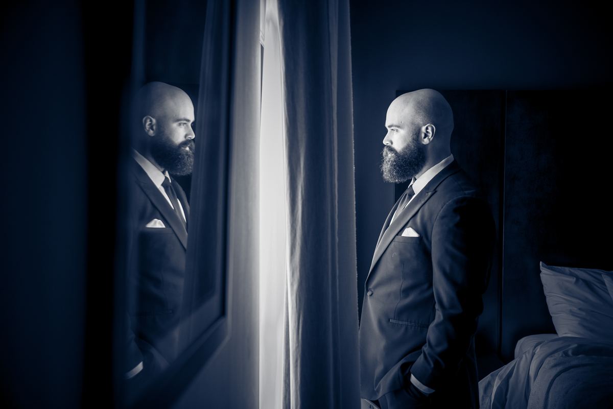 A bearded man in a suit stands by a partially covered mirror, illuminated by a vertical beam of light.