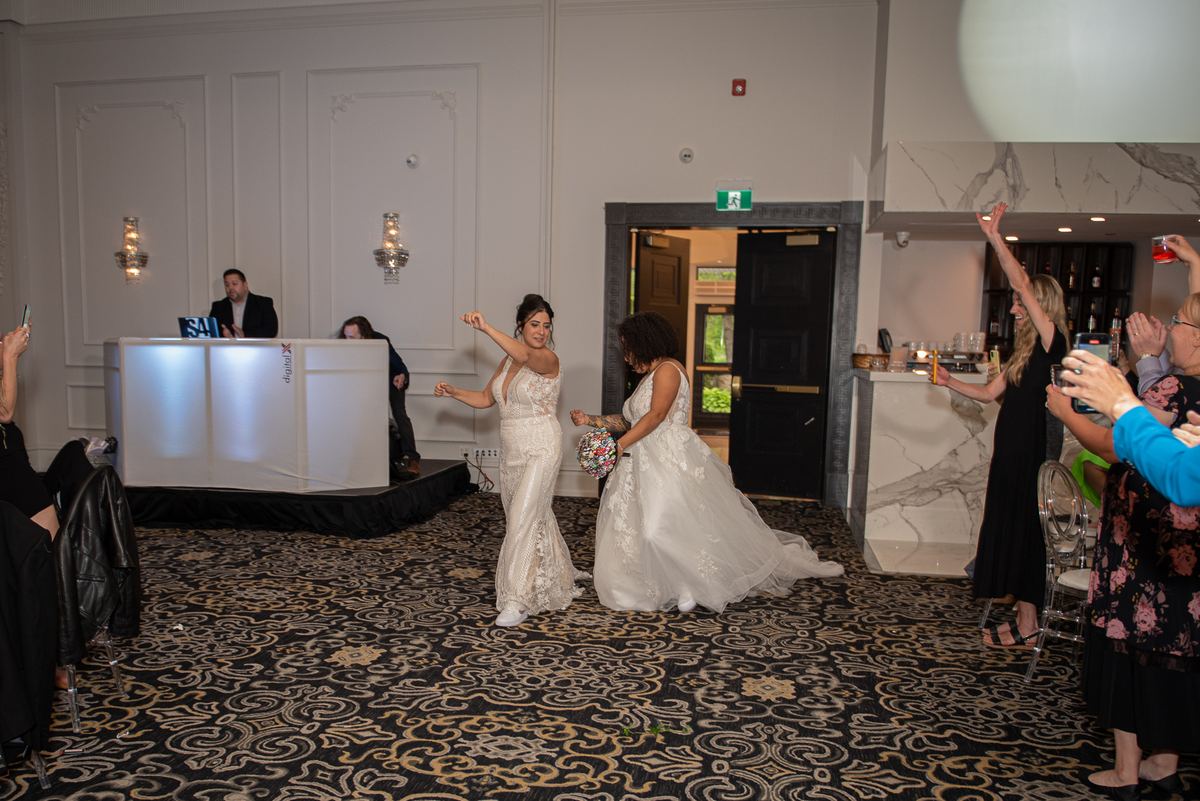 Two brides dance joyfully in a banquet hall, arms raised, surrounded by cheering guests and lit by wall sconces.
