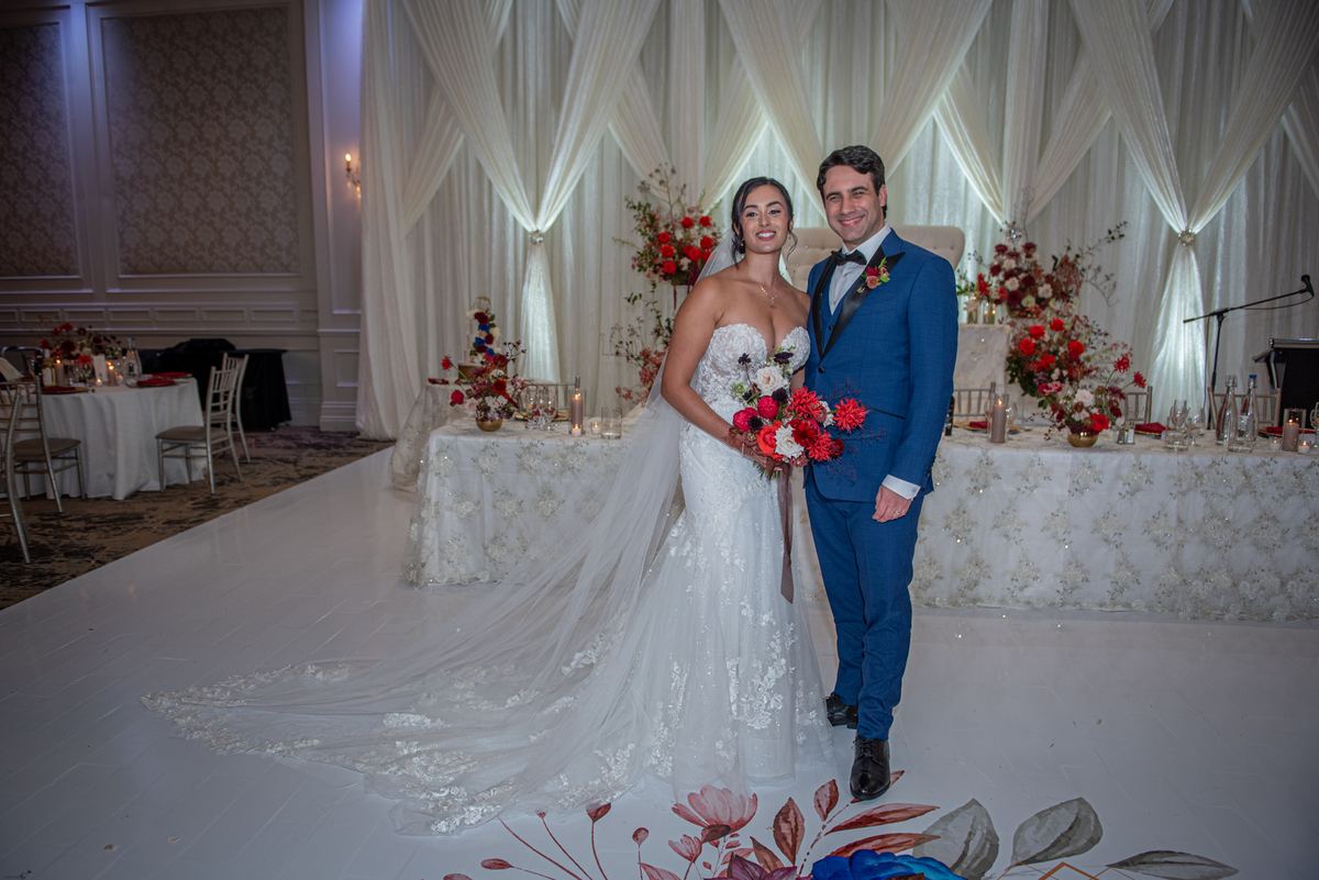 A smiling bride and groom stand on a decorated dance floor with white curtains and floral arrangements.