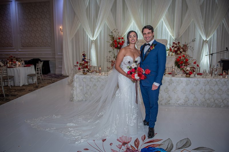 A smiling bride and groom stand on a decorated dance floor with white curtains and floral arrangements.