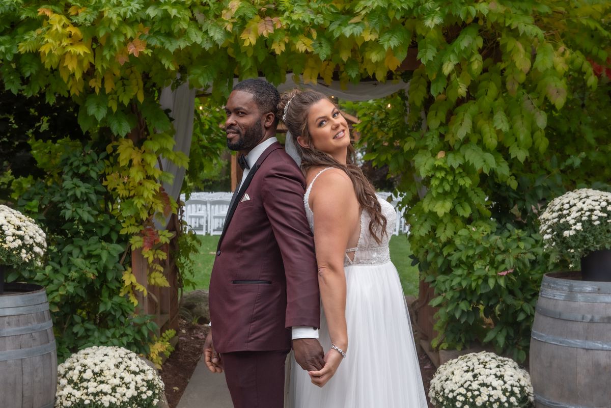 A smiling bride and groom hold hands under a green vine archway in a garden setting with wooden barrels and chrysanthemums.
