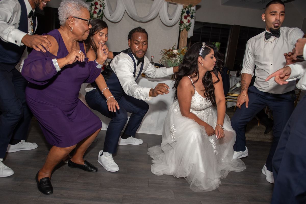 Guests including the bride dance energetically in a squatting style at a wedding reception, surrounded by smiling family.