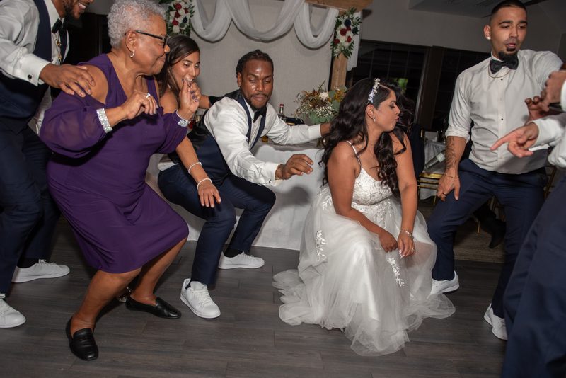Guests including the bride dance energetically in a squatting style at a wedding reception, surrounded by smiling family.