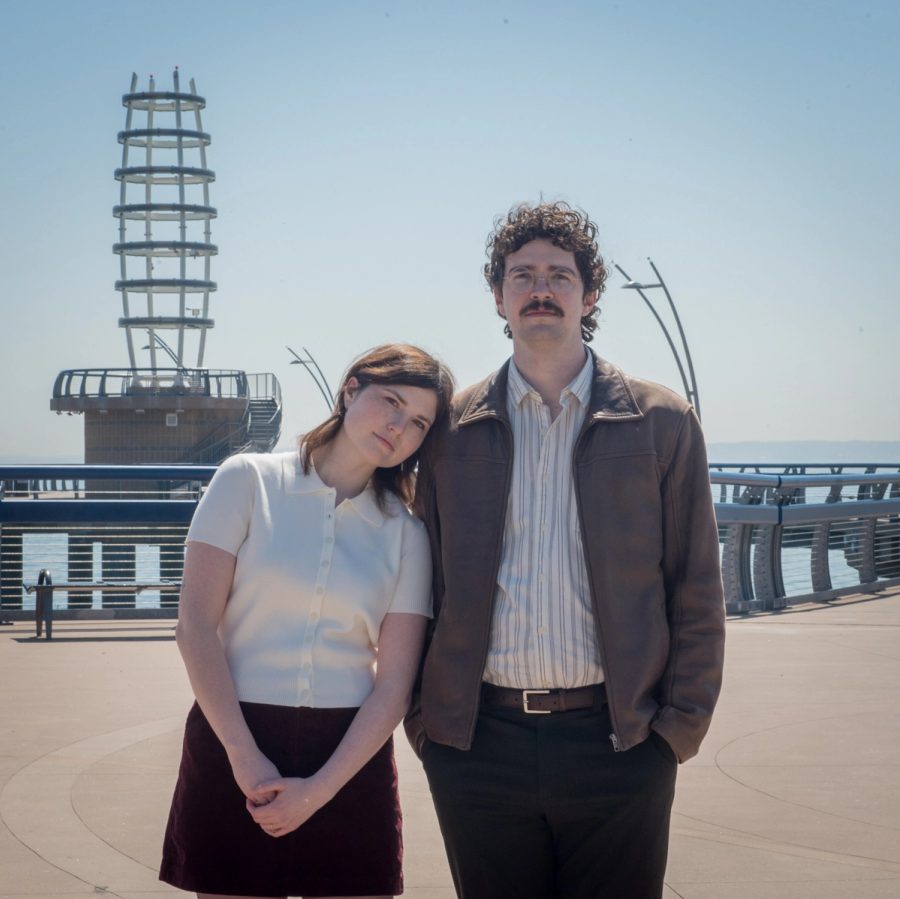 A couple stand together on a sunlit waterfront promenade, she resting her head on his shoulder, a slatted observation tower and the lake behind them.