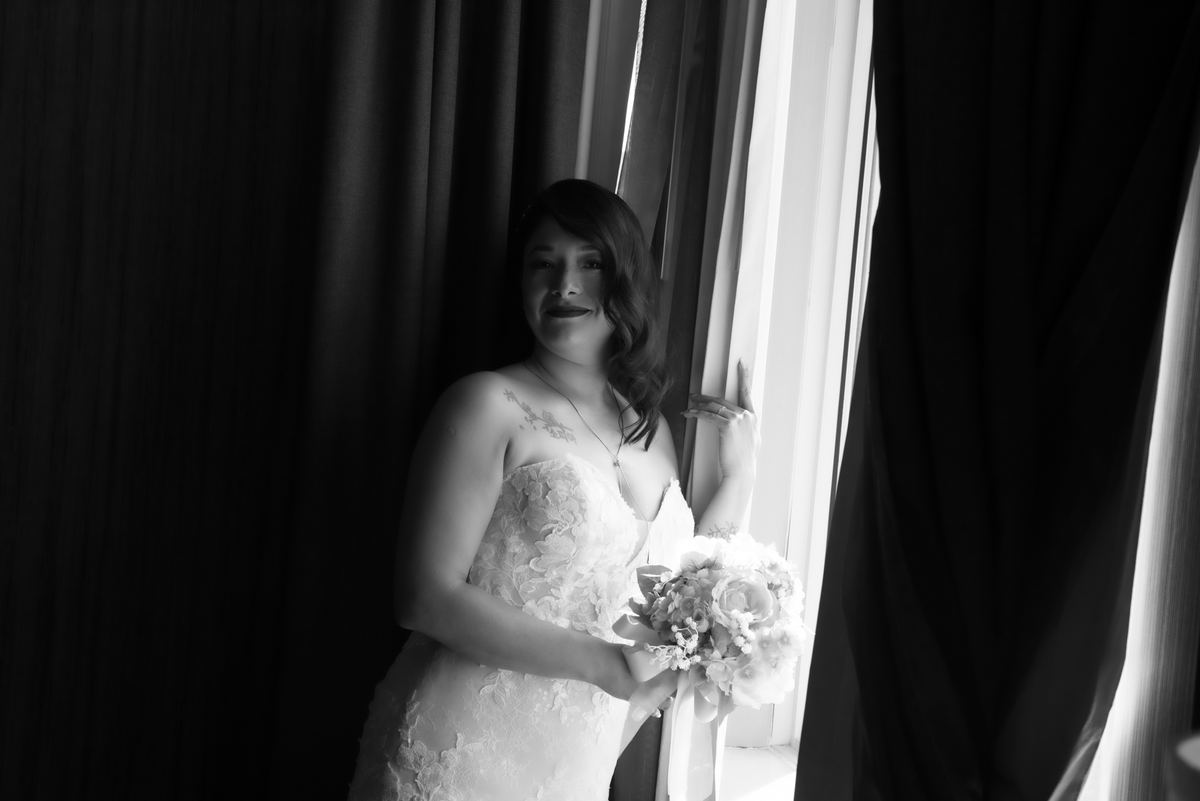 A bride in a glowing gown stands by a window, holding a bouquet and gazing toward the camera, framed by dark curtains.