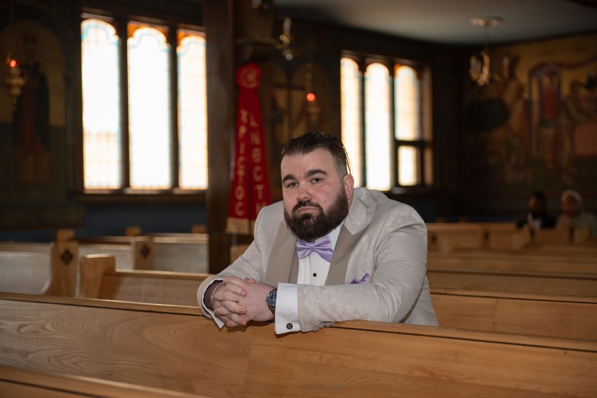 A man in a light tuxedo with a lavender bow tie stands by a pew, looking at the camera in a church with stained glass.