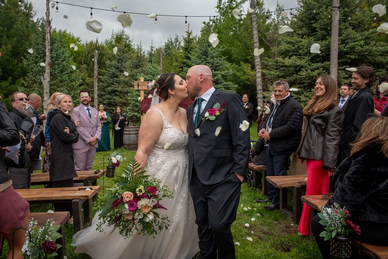 Anastasia and Adam share a tender kiss at their outdoor wedding as guests throw white petals into the air.