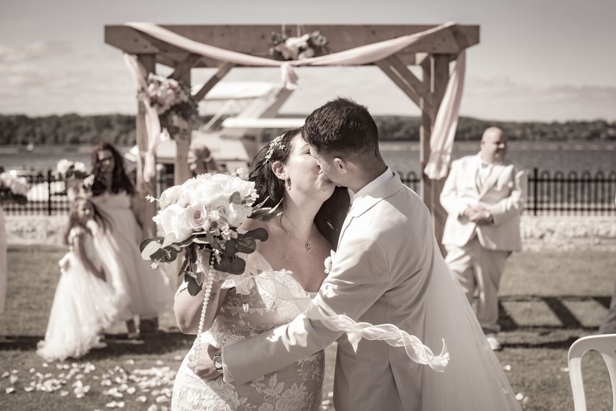 A bride and groom kiss beneath a decorated wooden arch by a lake, guests watching in the background.