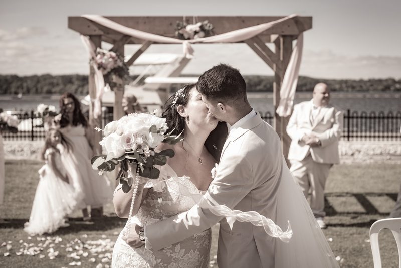 A bride and groom kiss beneath a decorated wooden arch by a lake, guests watching in the background.