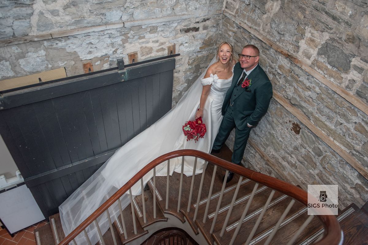 A bride and groom smile broadly at the camera while standing on a wooden staircase inside a stone-walled building.