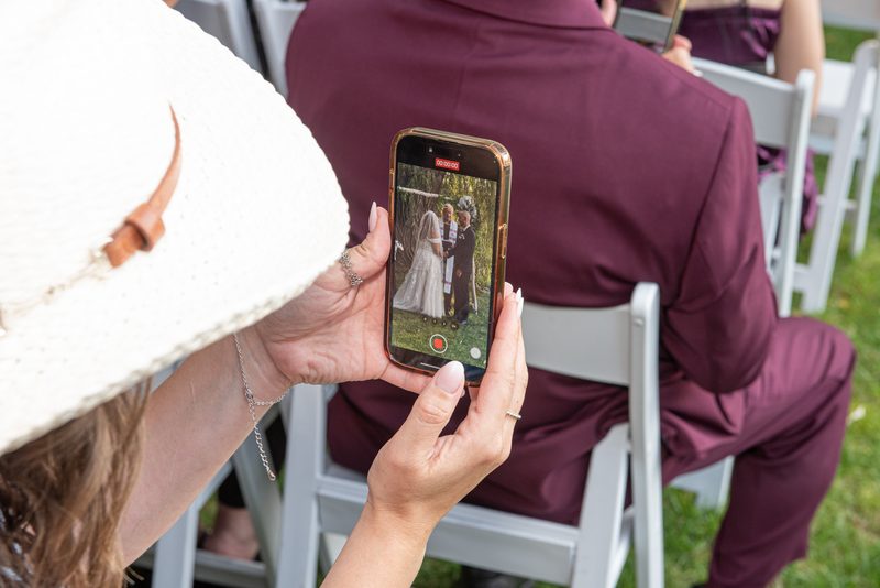 A woman records a wedding ceremony outdoors, showing the bride and groom under a wooden arch on a grassy lawn.