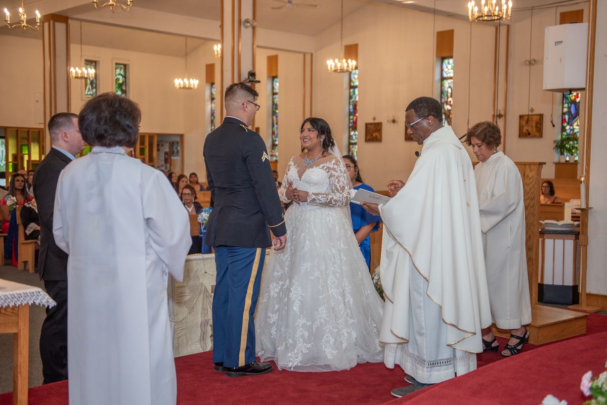 A bride in a lace gown and groom in military uniform smile, holding hands during their church wedding ceremony.