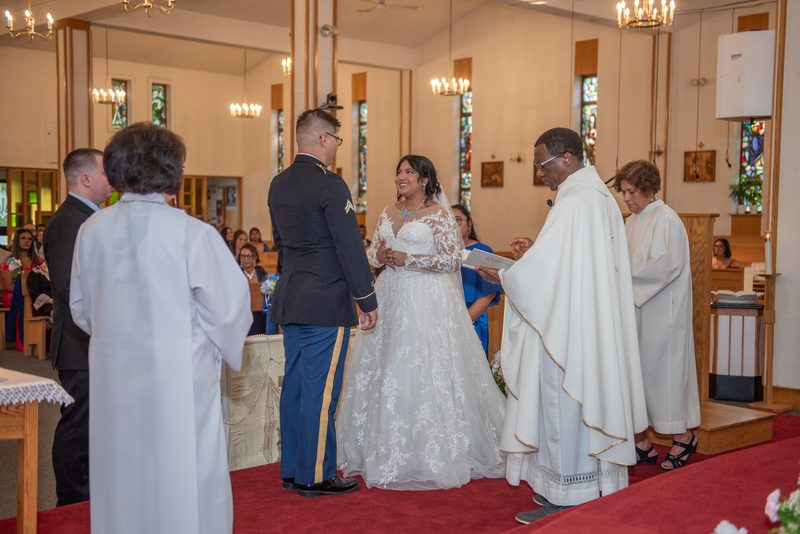 A bride in a lace gown and groom in military uniform smile, holding hands during their church wedding ceremony.