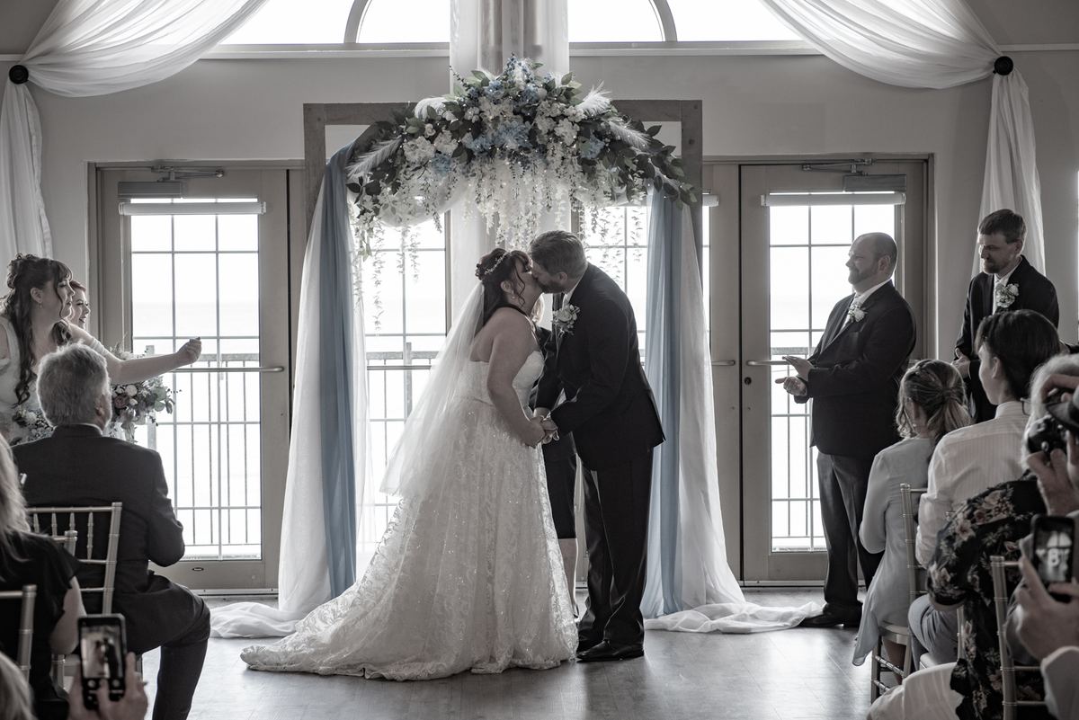 A bride and groom kiss beneath a floral arch at their wedding ceremony, guests watching from rows of seats.