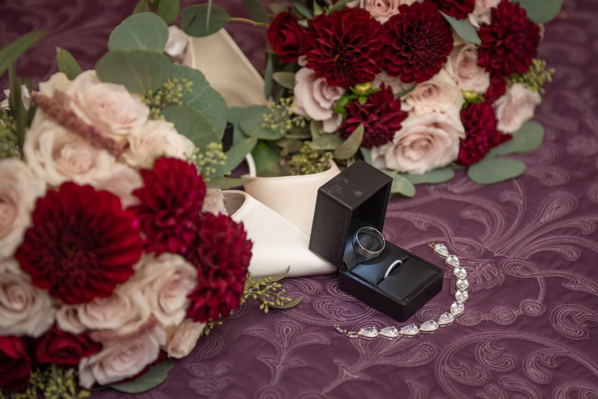A black ring box with engagement and wedding bands, a teardrop bracelet, a bridal bouquet of dahlias and roses.