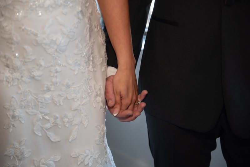 Bride and groom hold hands, fingers intertwined between lace veil and dark suit, in soft diffused light.