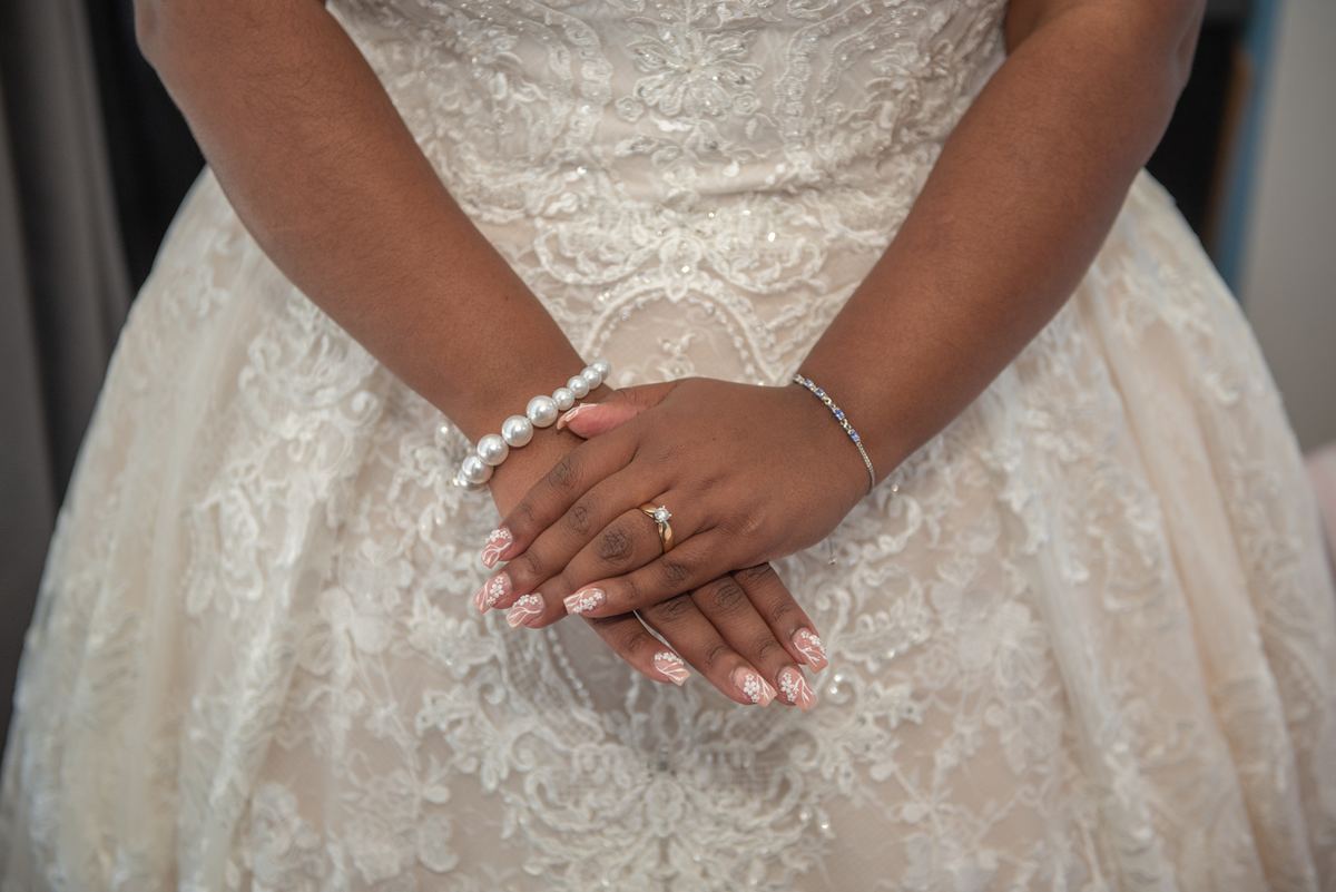 A bride's hands adorned with jewelry rest gently in front of her, the lace of her wedding gown visible.