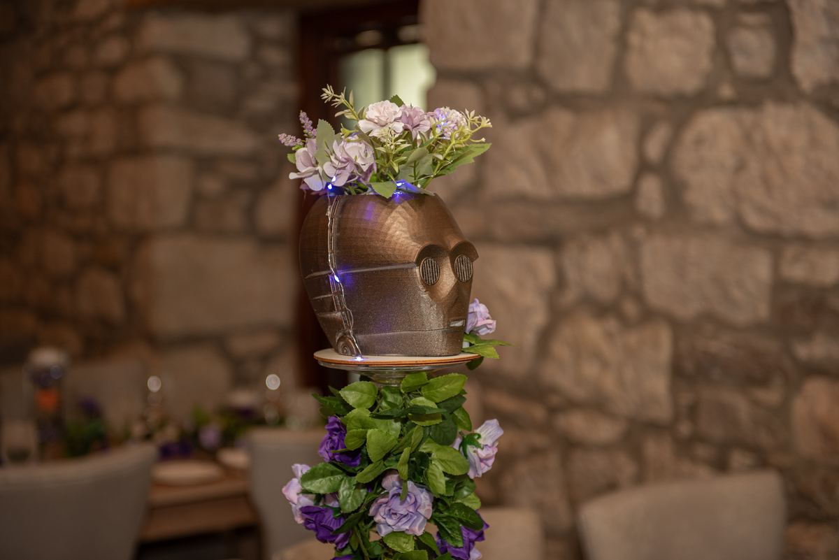 A metallic C-3PO head from Star Wars adorned with flowers and string lights sits on a pedestal surrounded by greenery.