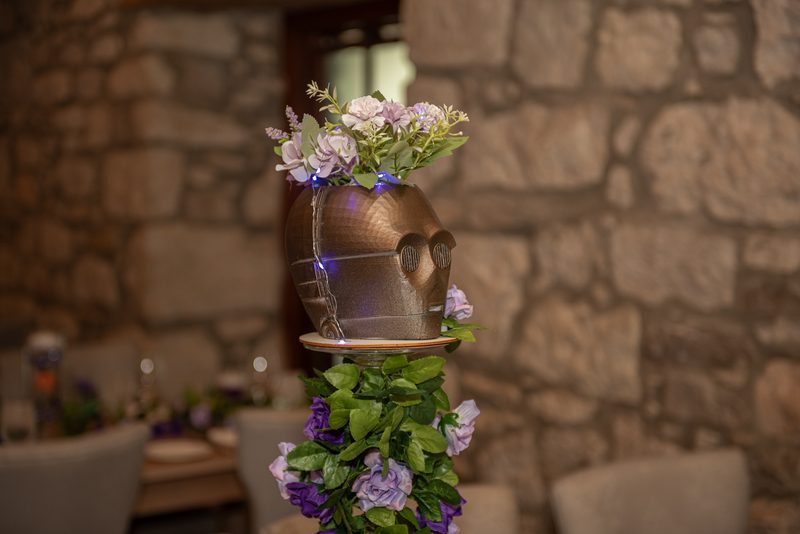 A metallic C-3PO head from Star Wars adorned with flowers and string lights sits on a pedestal surrounded by greenery.